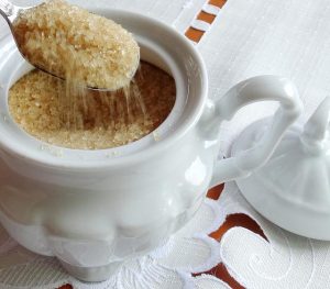 Açucareiro branco sobre uma mesa. A imagem mostra uma colher com açúcar saindo do açucareiro.